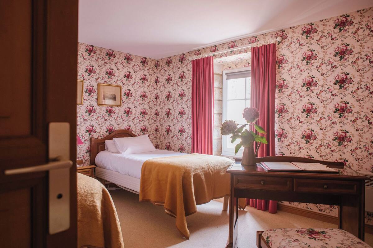 A bedroom with floral wallpaper, a bed with a white comforter and a mustard-colored blanket, a wooden desk with a vase of flowers, and a window with pink curtains.