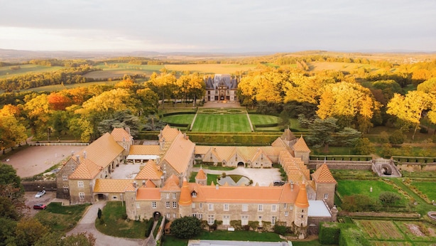 Un edificio grande e histórico con múltiples torres y un patio central, rodeado de campos extensos y follaje otoñal.