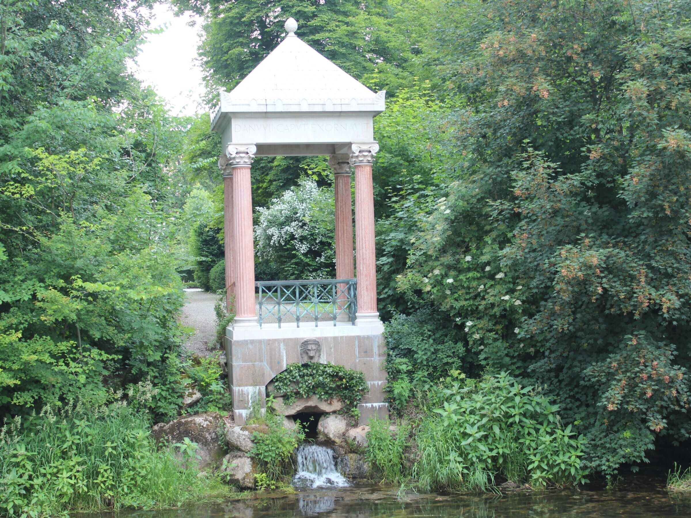 Een stenen kiosk met zuilen en een dak, omgeven door weelderige begroeiing en een kleine waterval.