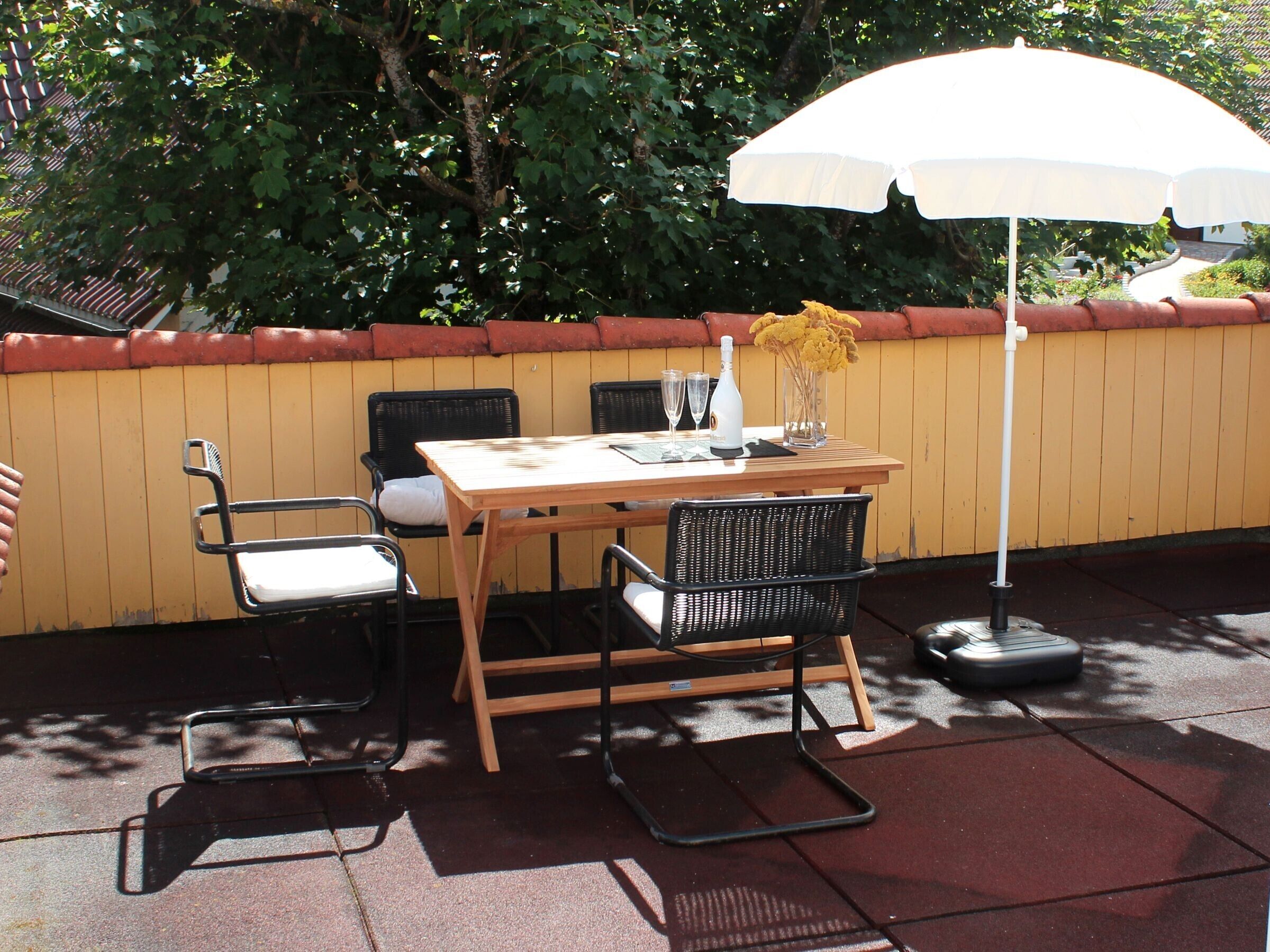 Una terrazza esterna con tavolo di legno, sedie nere, ombrellone bianco e un vaso con fiori gialli.