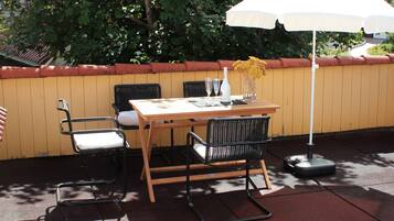 Una terrazza esterna con tavolo di legno, sedie nere, ombrellone bianco e un vaso con fiori gialli.