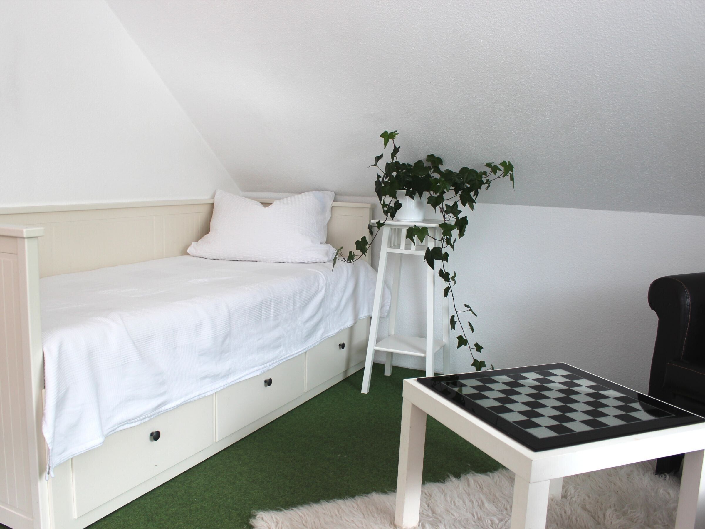 A bedroom with a bed, a small table with a chessboard, a white chair, and a plant.