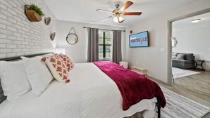 Un dormitorio moderno con pared de ladrillo blanco, ventilador de techo, cama con ropa de cama blanca y roja, televisor en la pared y un sofá gris en la habitación contigua.