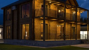 Een houten chalet met twee verdiepingen, een balkon, grote ramen en een goed verlichte buitenkant.