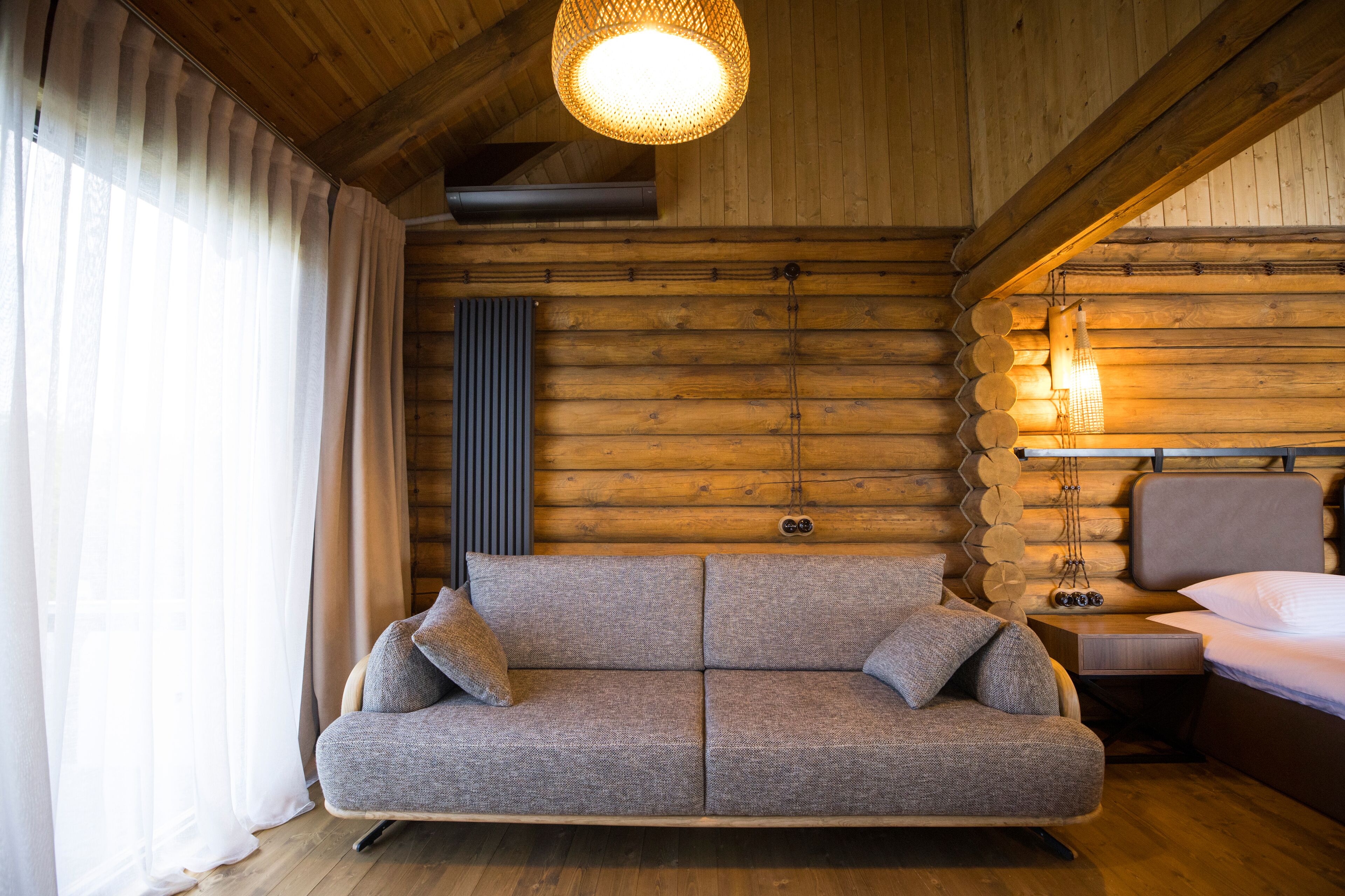 A log cabin interior with a sofa, a bed, a hanging light fixture, and a window with curtains.