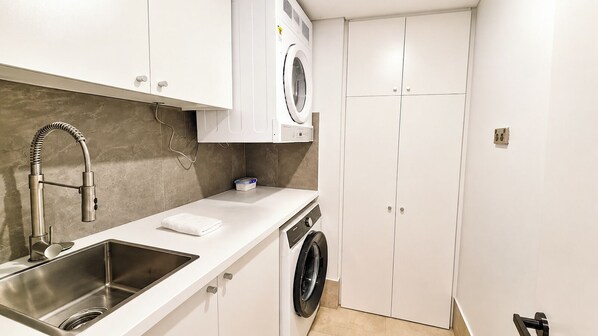 Cocina compacta con lavadora integrada, pileta de acero inoxidable y muebles bajos blancos.
