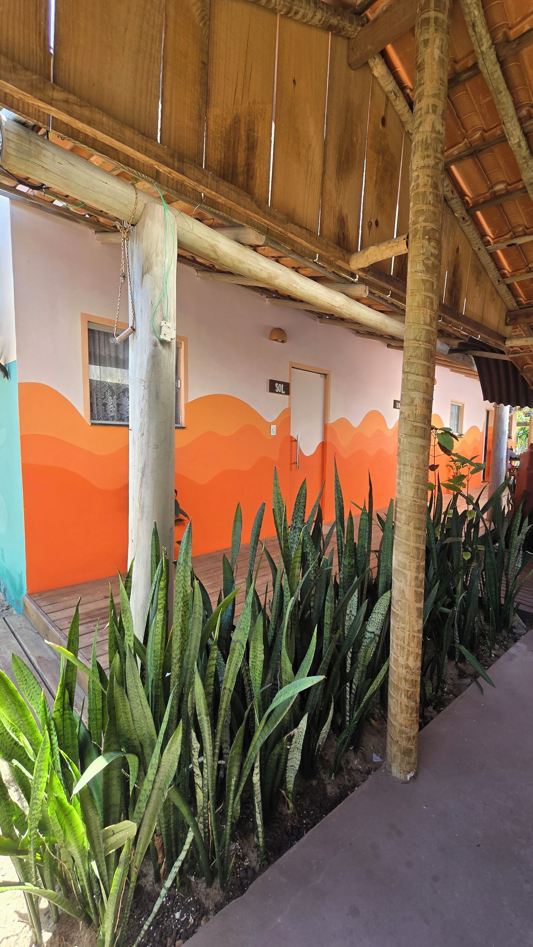 Un espacio al aire libre techado, con techo de madera, pared decorada con un mural de olas y una fila de plantas verdes.