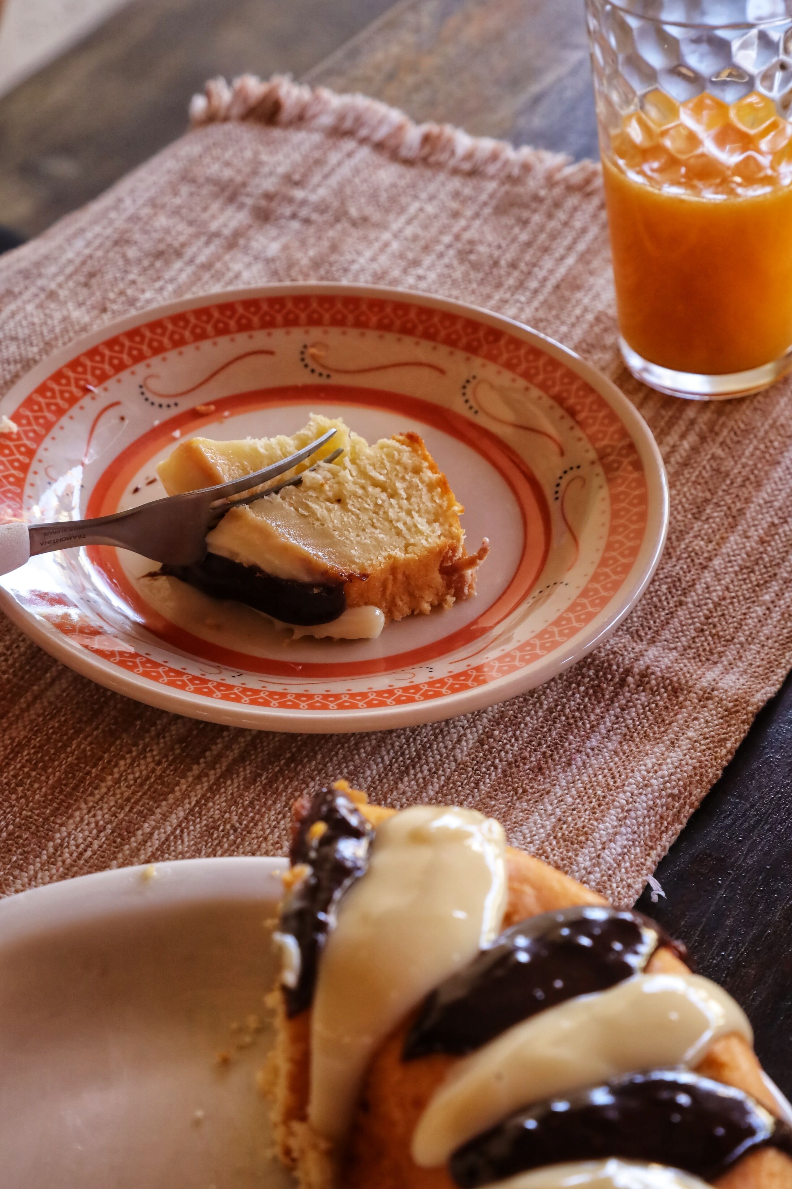 A slice of cake with a fork on a patterned plate, a glass of orange juice, and a napkin on a table.