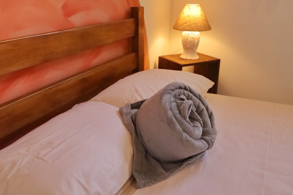 A neatly made bed with a rolled towel, a wooden headboard, a nightstand with a lamp, and a wall with a curtain.