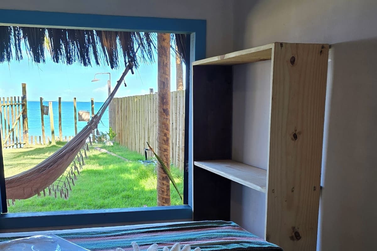 A bed with a view of a hammock and a wooden fence, a window, and a shelf unit.