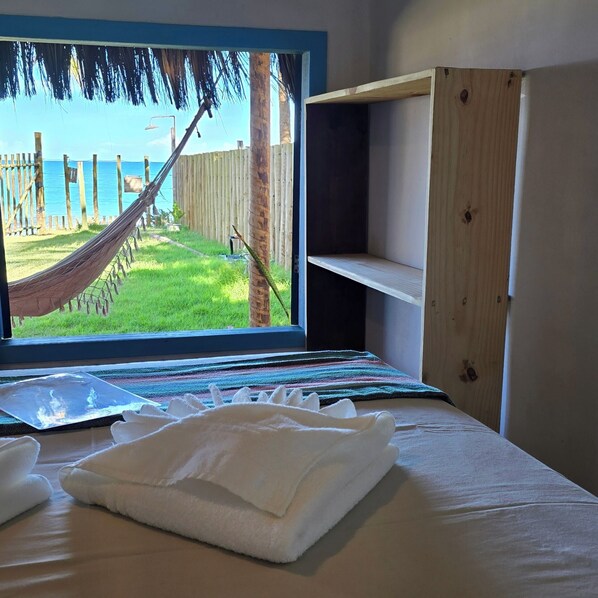 A bed with a view of a hammock and a wooden fence, a window, and a shelf unit. - Vila Ohana Beira Mar (Porto Seguro)