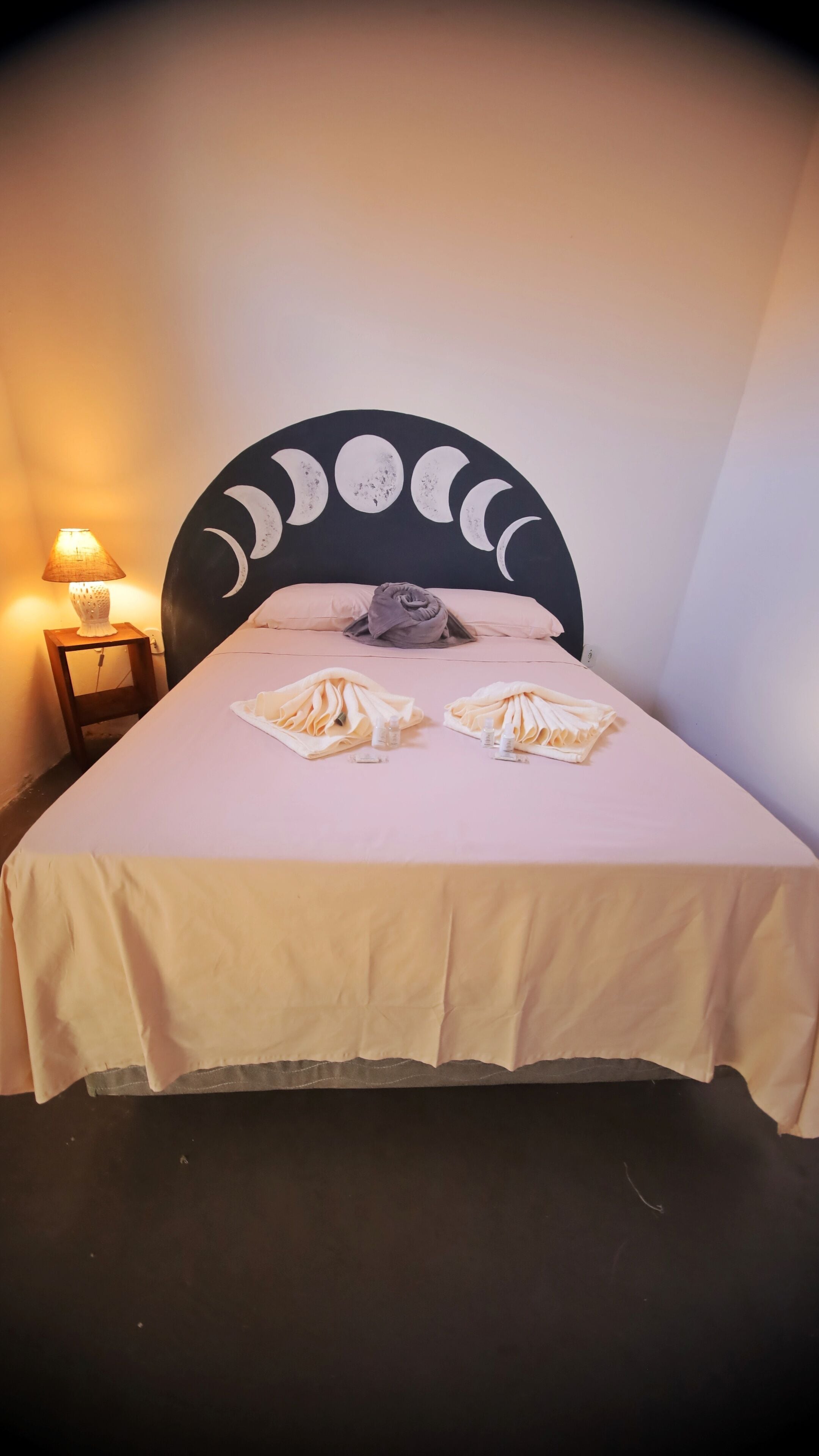 Un lit bien fait, avec une tête de lit ornée de motifs représentant les phases de la lune, une lampe de chevet et des serviettes soigneusement disposées sur le lit.