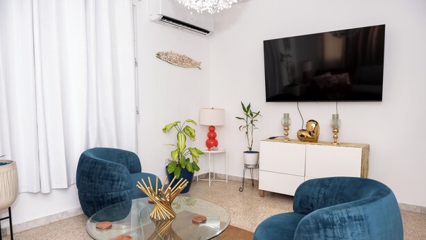 A modern living room with a glass coffee table, blue armchairs, a white cabinet, and a flat-screen TV mounted on the wall. - Cozy Home Close to Beaches (Cabo Rojo)