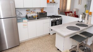 A compact kitchen with white cabinets, a stainless steel refrigerator, a microwave, a gas stove, and a small island with stools. - Cozy Home Close to Beaches (Cabo Rojo)