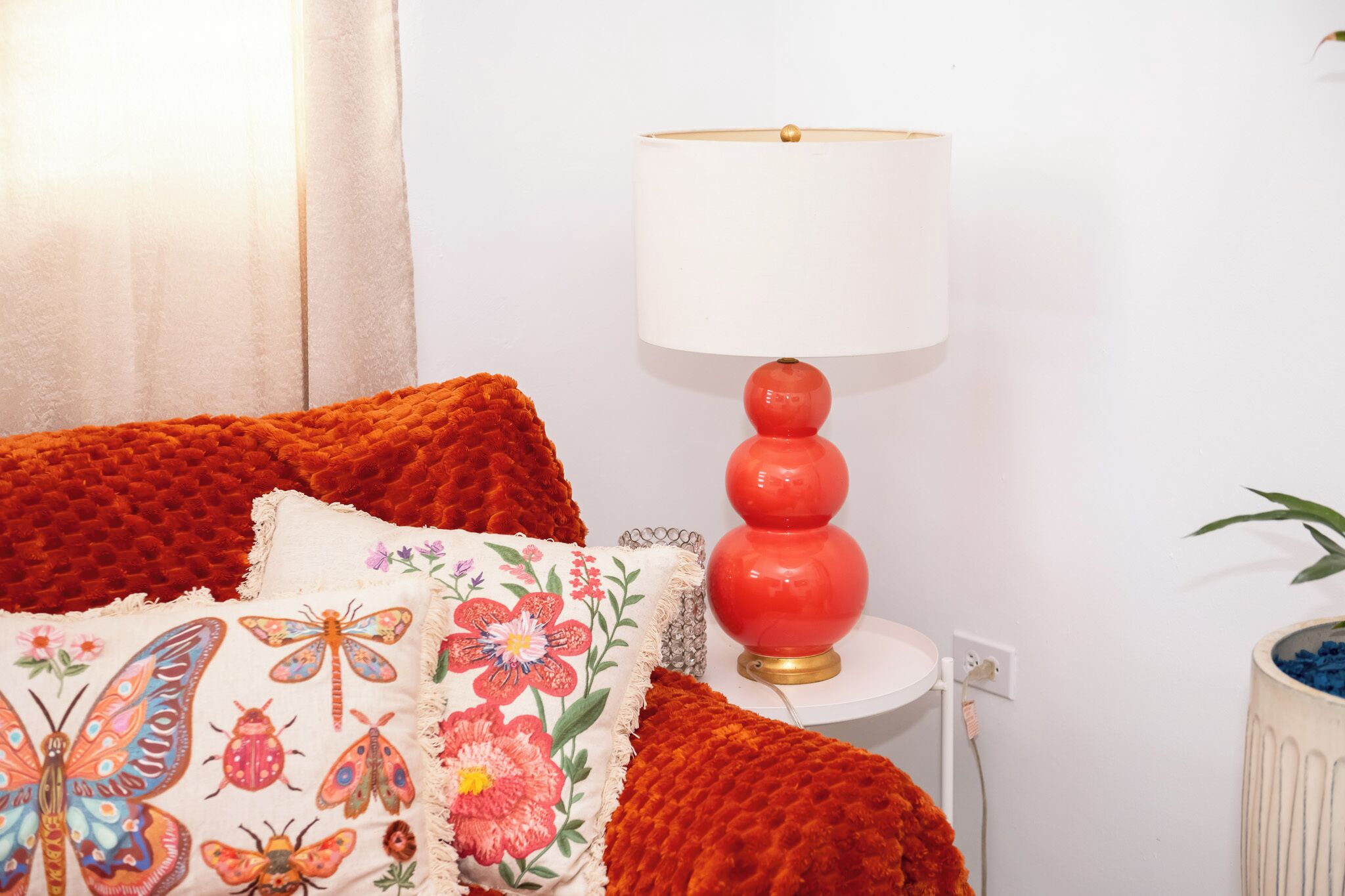 Ein Zimmer mit einer rot gemusterten Bettdecke, einem weißen Kissen mit aufgestickten Blumen und Insekten sowie einer roten Lampe mit weißem Lampenschirm auf einem weißen Nachttisch.