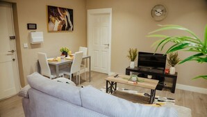 A modern living room with a sofa, a dining table with chairs, a TV on a black stand, and a clock on the wall. - 1BR Retreat l Great Value (West Midlands)