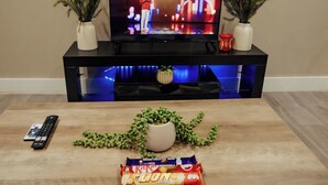 A living room with a TV on a black stand, two potted plants, a remote control, a plant on the floor, and a snack package. - 1BR Retreat l Great Value (West Midlands)