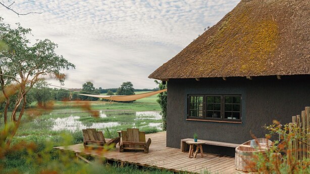 A thatched-roof house with a wooden deck, two wooden chairs, a small table, and a hammock. - Cottage "Am Wald" links (Bissee)