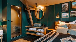 A modern bathroom with a glass shower, wooden shelves with towels, a wooden bench, and a bed with pillows and a blanket.
