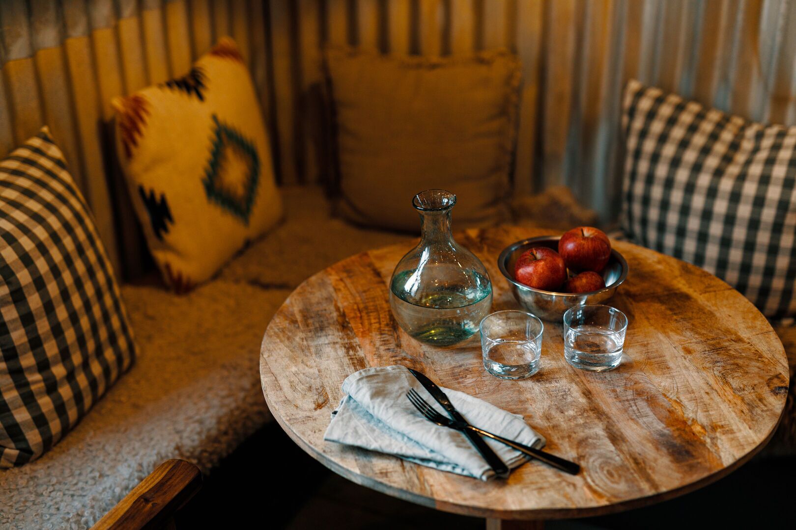 A round wooden table with a glass bottle, a bowl of apples, two shot glasses, and a folded napkin with cutlery.