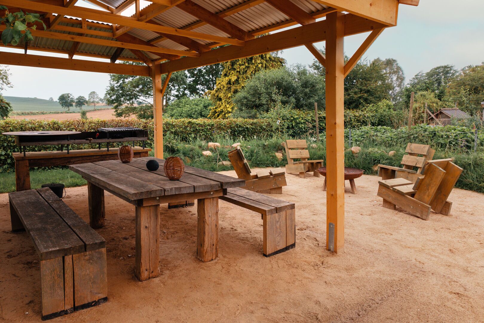 An outdoor patio with a wooden table and benches, surrounded by greenery and a barbecue grill.