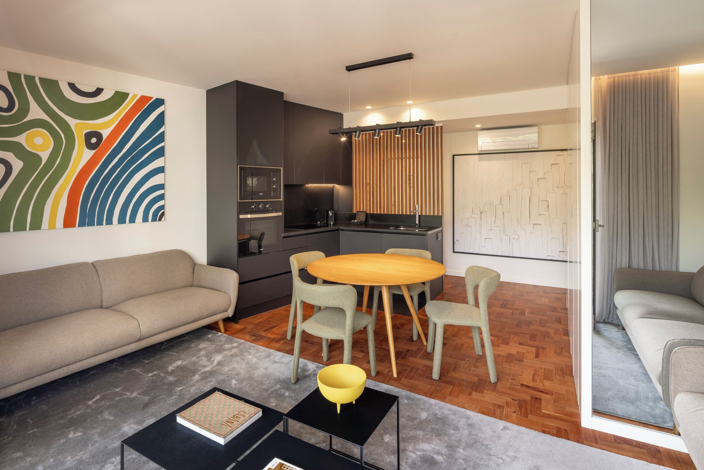 A modern living room with a sofa, a dining table with chairs, a kitchen area, and a large abstract painting on the wall.