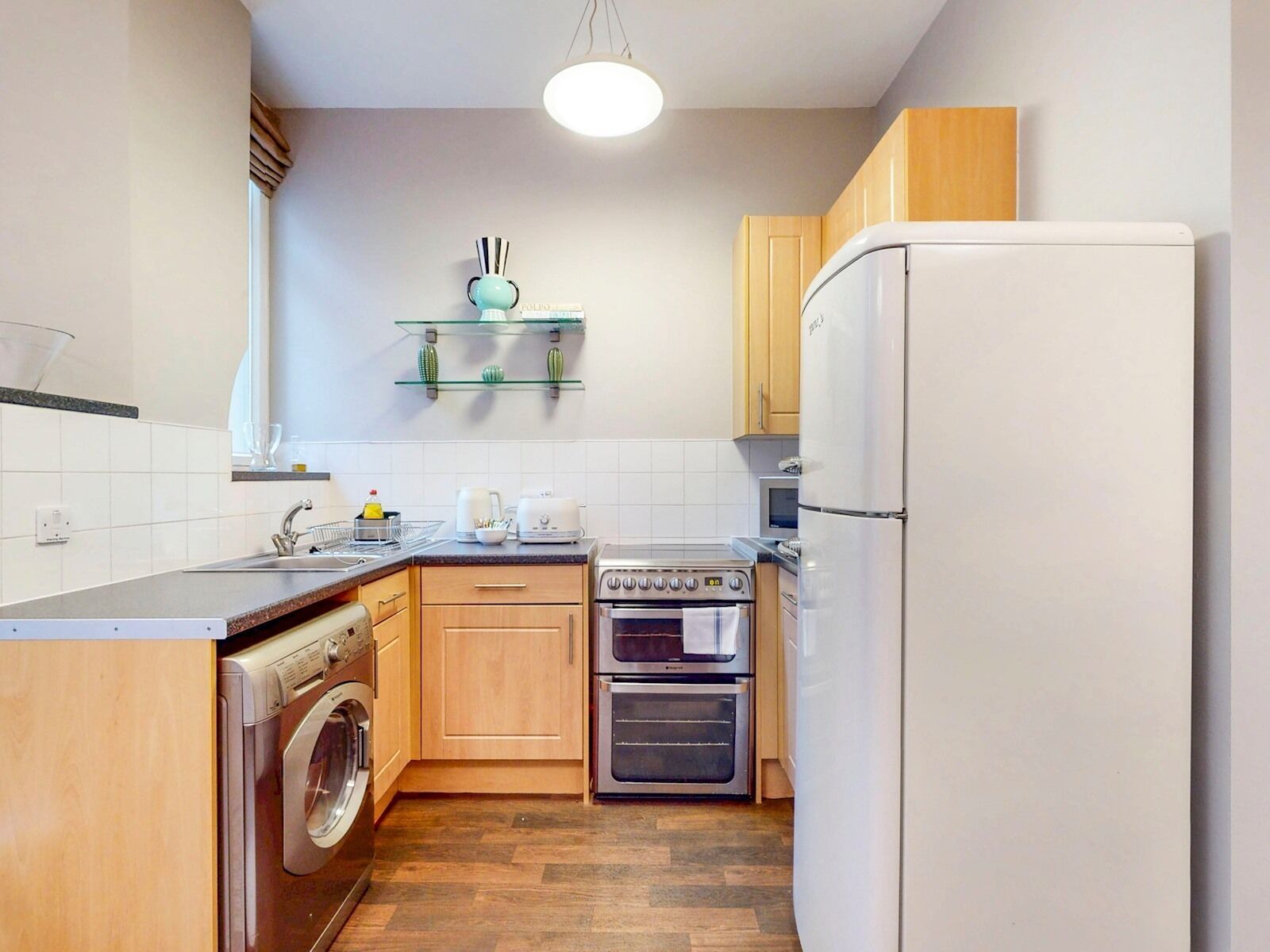 A compact kitchen with a washing machine, a double oven, a refrigerator, and a small shelf with decorative items.