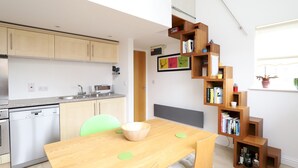 Una cocina moderna con lavavajillas, un fregadero y un microondas. Hay una mesa de comedor de madera con sillas y una pequeña estantería con libros y objetos decorativos.