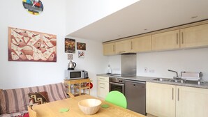 Una cocina compacta con armarios de madera, microondas, tostadora y lavavajillas. Hay una mesa de comedor de madera con un tazón y manteles individuales verdes. Un cuadro abstracto colorido y un reloj están en la pared.