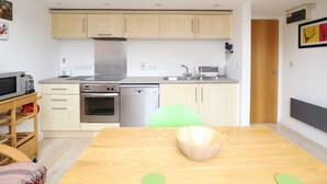 Cocina compacta con microondas, tostadora, horno y lavavajillas. Hay una mesa de madera con un tazón y una silla.