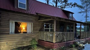 Une cabane en bois avec un toit rouge, une véranda dotée d’une balustrade en bois et une fenêtre derrière laquelle on aperçoit une lampe allumée.