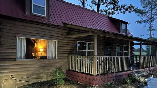 Eine Holzhütte mit rotem Dach, einer Veranda mit Holzgeländer und einem Fenster mit brennender Lampe im Inneren.