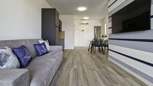 A modern living room with a grey sofa, patterned cushions, a dining area with black chairs, and a TV mounted on the wall. - Bright Studio Apartment in Bibione (San Michele al Tagliamento)