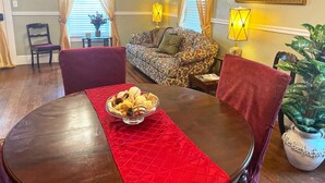 A dining area with a round table, red placemat, and a bowl of food. There is a sofa with patterned cushions, a chair, and a potted plant. - Two Bedroom House Close to Local Favorites (West Lafayette)