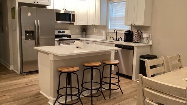 Una cocina moderna con gabinetes blancos, un refrigerador de acero inoxidable, un microondas, un horno y una cafetera. Hay tres taburetes de madera y una mesa de comedor con sillas.