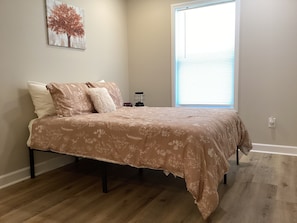 A bedroom with a bed, a nightstand, a window with blinds, and a wall-mounted artwork.