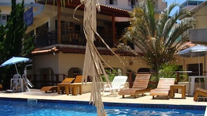 Une piscine entourée de chaises longues et de parasols, le tout bordé d’un bâtiment doté de balcons et de palmiers.