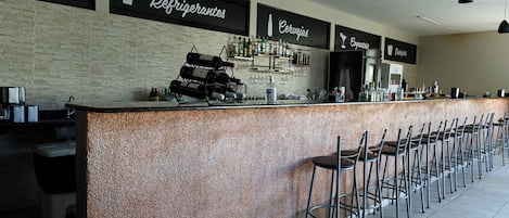Una zona de bar con una barra larga, varios taburetes y estantes llenos de botellas y vasos.