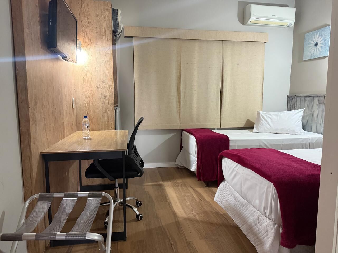 Een compacte hotelkamer met een houten bureau, een stoel, twee eenpersoonsbedden met rode dekens, een tafeltje met een waterfles en een luchtverkoeler aan de muur.