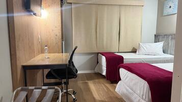 Habitación de hotel compacta con un escritorio de madera, una silla, dos camas individuales con mantas rojas, una mesita con una botella de agua y un aire acondicionado instalado en la pared.