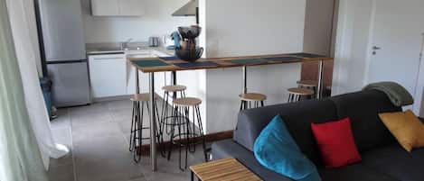 Un espace de vie compact comprenant un coin cuisine, un espace repas avec des tabourets, un canapé avec des coussins et une petite table en bois.