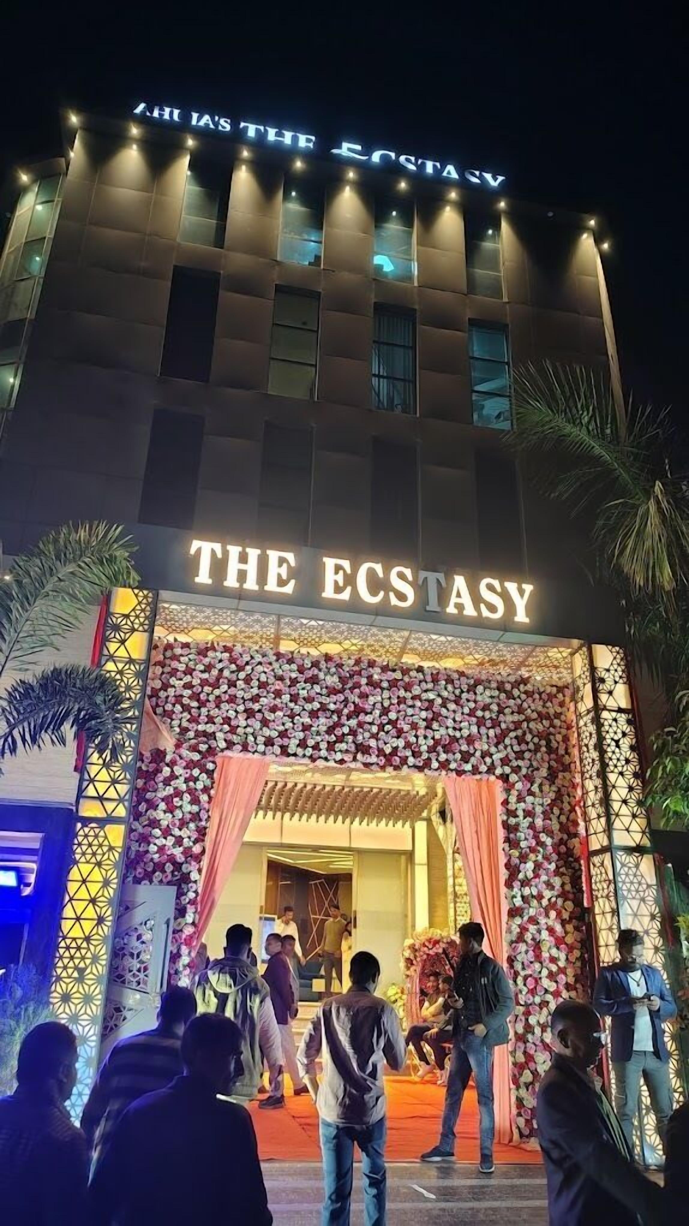 A building with a sign reading "THE ECSTASY" at the entrance, surrounded by decorative floral arrangements and palm trees.