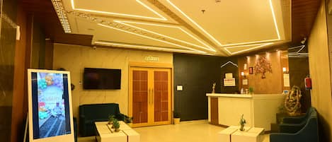 A hotel lobby with a reception desk, two sofas, a TV screen, and a decorative wall.