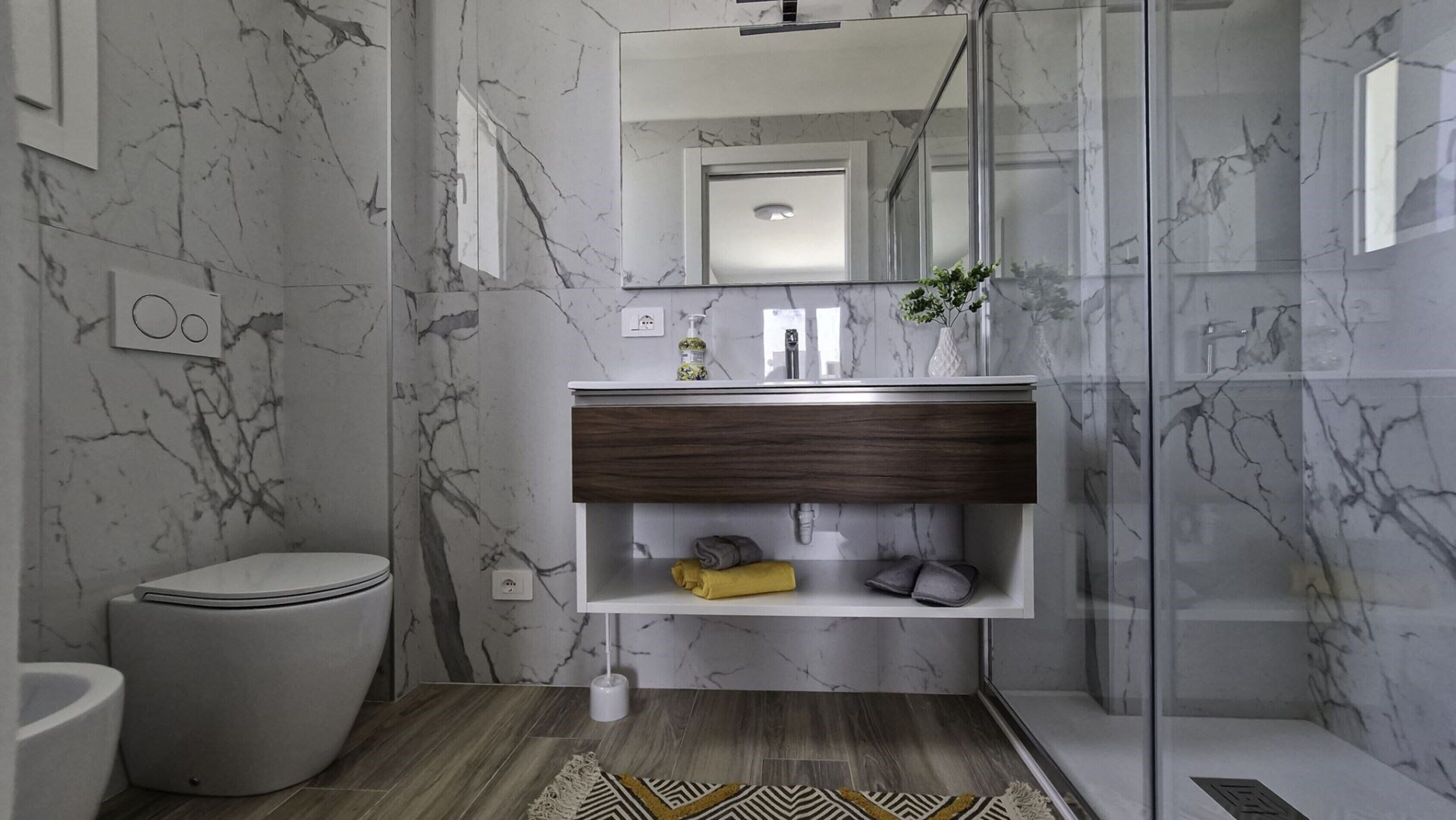 A modern bathroom with a marble wall, a white toilet, a wooden vanity, and a glass shower enclosure.