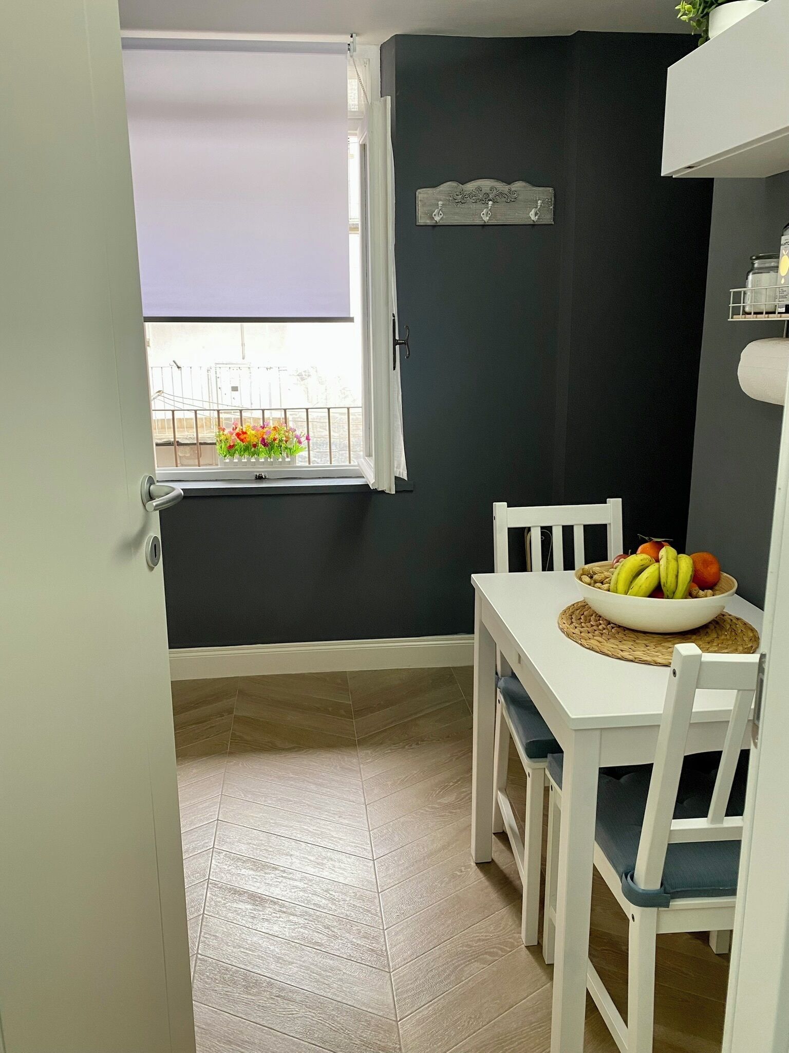 Uma pequena área de refeições com uma mesa e cadeiras brancas, uma tigela de frutas e uma janela com vista para uma varanda.