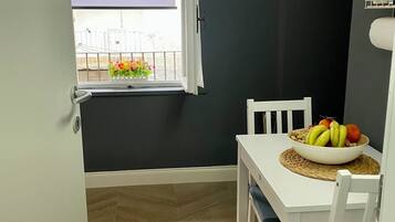 Uma pequena área de refeições com uma mesa e cadeiras brancas, uma tigela de frutas e uma janela com vista para uma varanda.