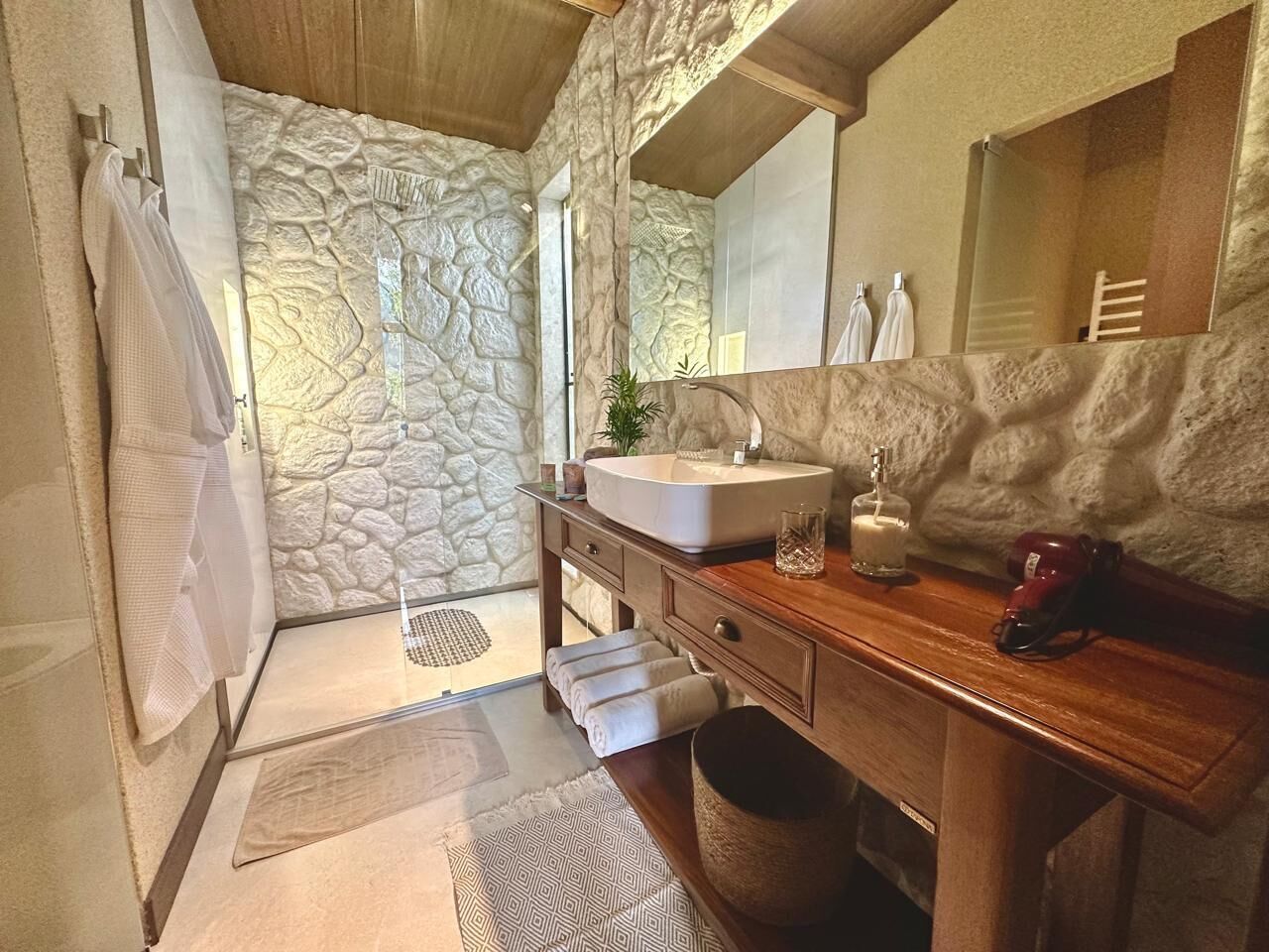 Un baño con pared de piedra, tocador de madera, lavamanos y espejo.