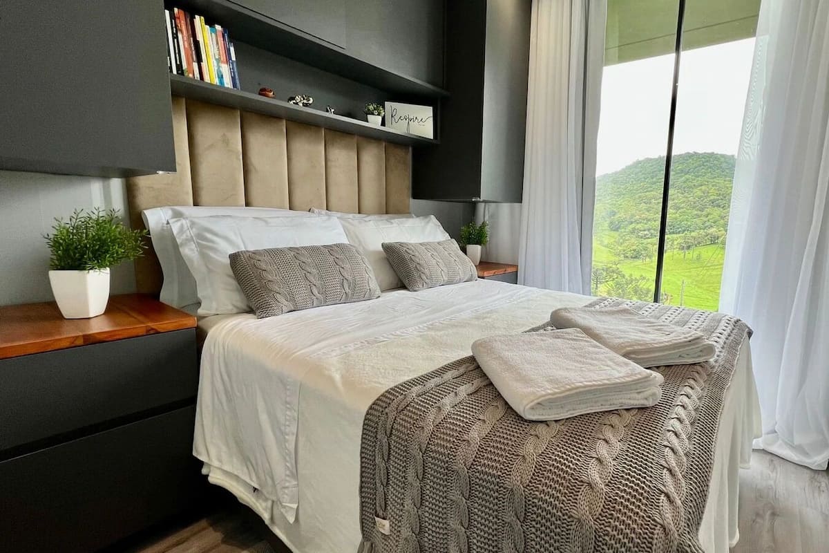Een moderne slaapkamer met een bed, een nachtkastje met een plant, een plank met boeken en uitzicht op groen buiten.