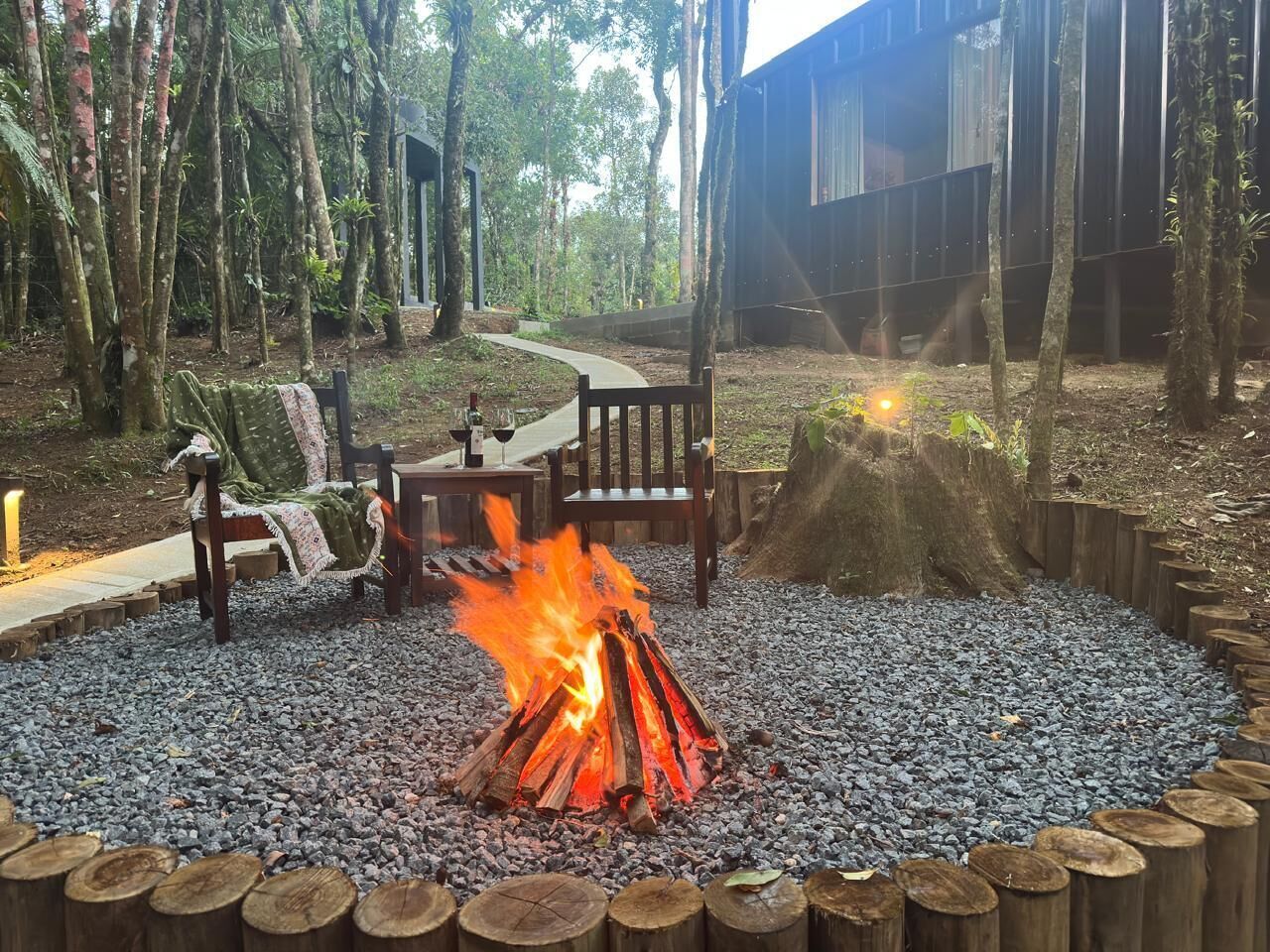 A fire pit surrounded by a circular arrangement of wooden logs and pebbles, with chairs and a table nearby.