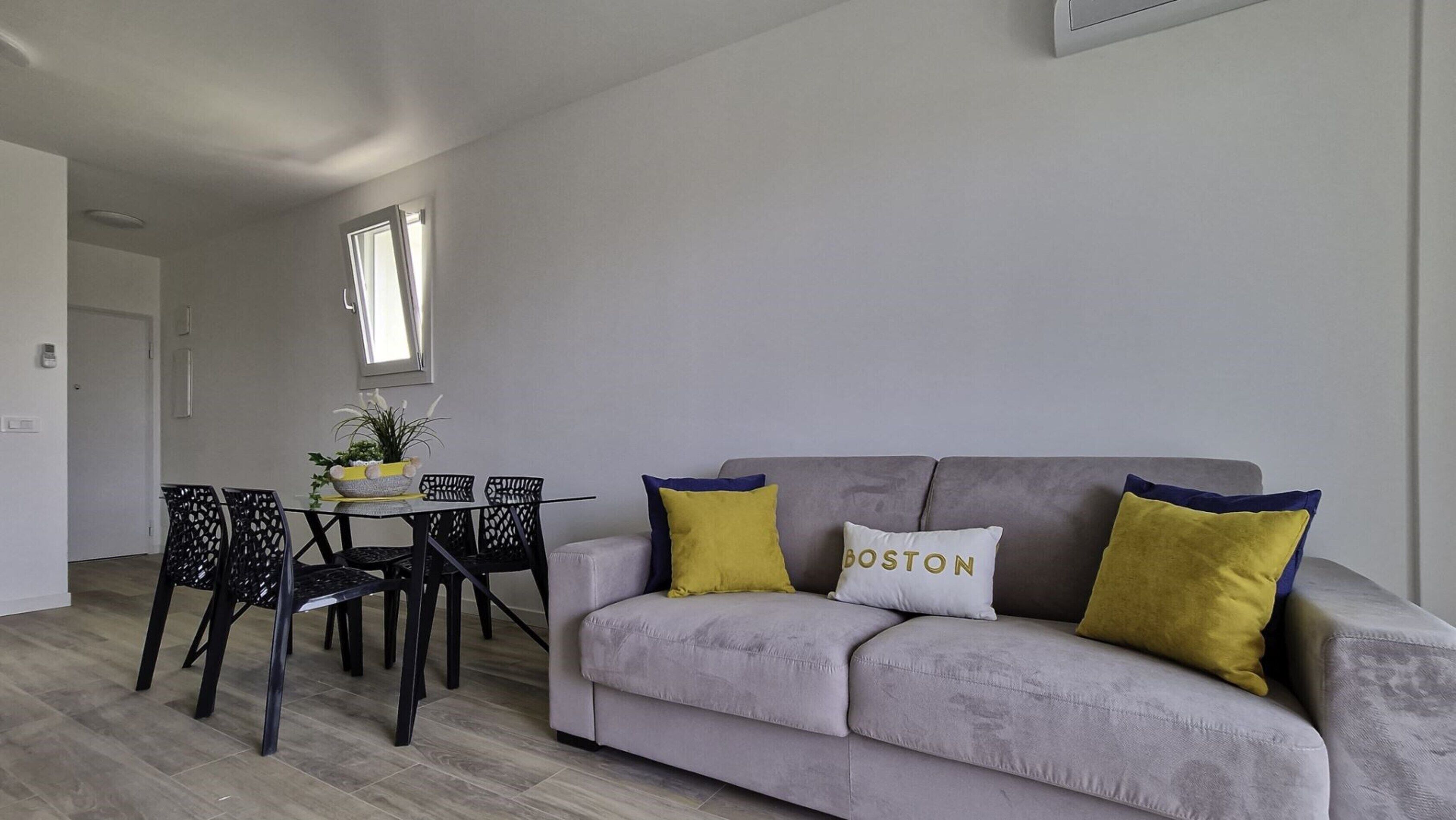 Olohuoneessa on harmaa sohva, kaksi keltaista koristetyynyä, valkoinen tyyny, jossa on teksti ”BOSTON”, pieni pöytä tuoleineen ja ikkuna, jossa on sälekaihtimet.